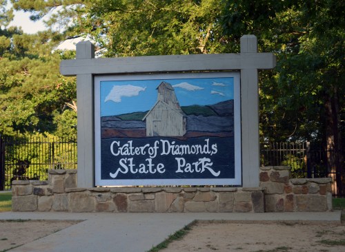 Sign for Crater of Diamonds State Park featuring a painted depiction of a diamond mine and surrounding landscape.