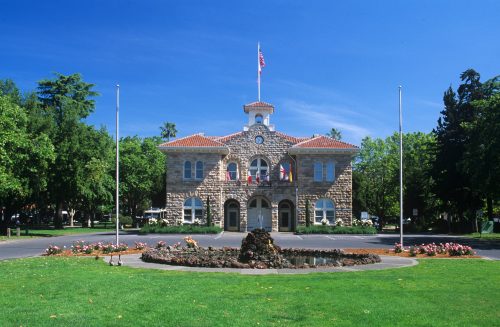 Historic stone building with a clock tower, surrounded by green grass and trees, featuring a fountain in the foreground.