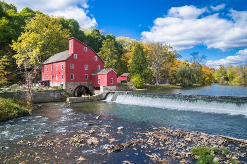 A red historic mill beside a river, with colorful trees and a waterfall under a blue sky with fluffy clouds.