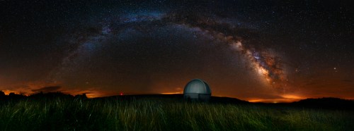 A panoramic view of the Milky Way arching over a grassy field and an observatory dome under a starry night sky.