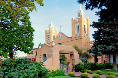 A charming adobe-style church with twin towers, surrounded by lush greenery and colorful flowers.