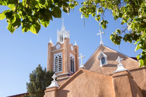 A church with a tall steeple and multiple crosses, framed by green leaves against a clear blue sky.