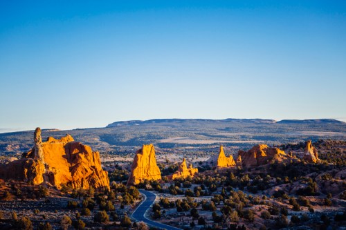 A scenic view of red rock formations under a clear blue sky, with a winding road and distant mountains in the background.