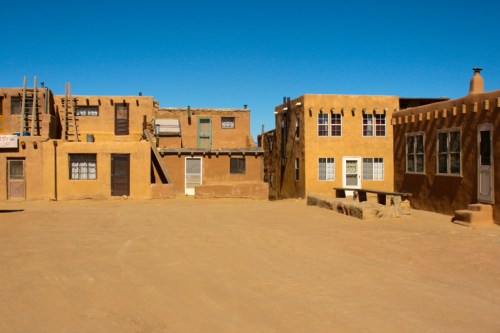 Adobe-style buildings in a desert landscape under a clear blue sky, with a sandy courtyard in the foreground.