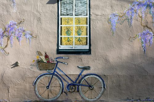 A blue bicycle with a basket rests against a textured pink wall adorned with purple flowers and a window.