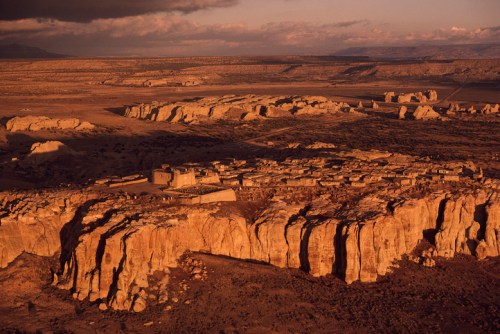 Aerial view of a desert landscape with cliffs and a small village nestled among the rocks at sunset.