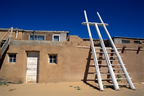 A traditional adobe building with a white ladder leaning against it, set against a clear blue sky.