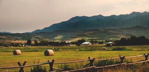 A serene landscape featuring rolling hills, mountains, and hay bales in a green field, with a wooden fence in the foreground.