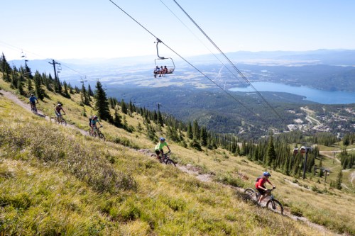 Mountain bikers ride down a grassy slope with a lake in the background, while a chairlift carries passengers above.