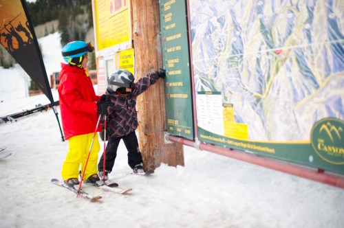 Two children in ski gear study a trail map at a snowy ski resort, with mountains in the background.