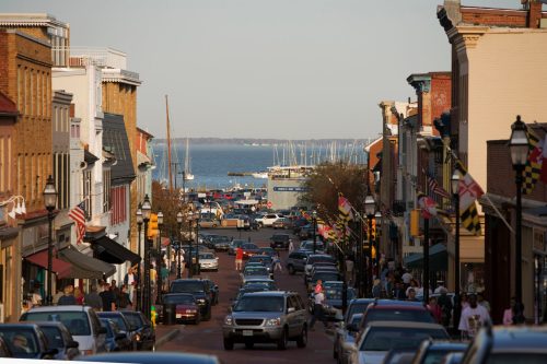 A bustling street lined with shops and cars, leading to a marina with sailboats and a clear blue sky.