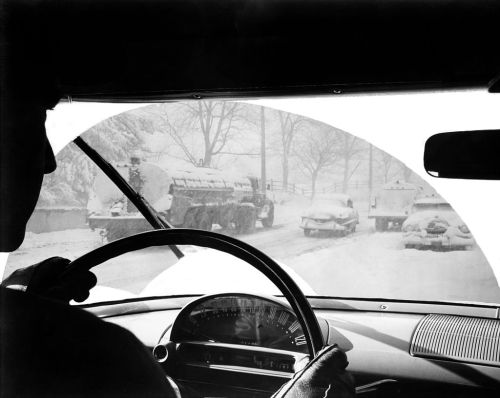 A driver views a snowy road through a car windshield, with snow-covered vehicles and trees in the background.
