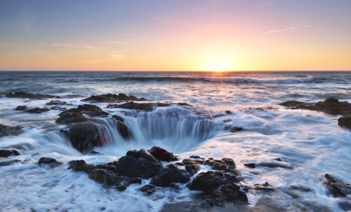 A serene sunset over the ocean, with waves cascading over rocky formations.