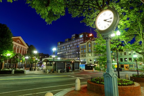 A city street at night featuring a vintage clock, trees, and modern buildings illuminated by streetlights.