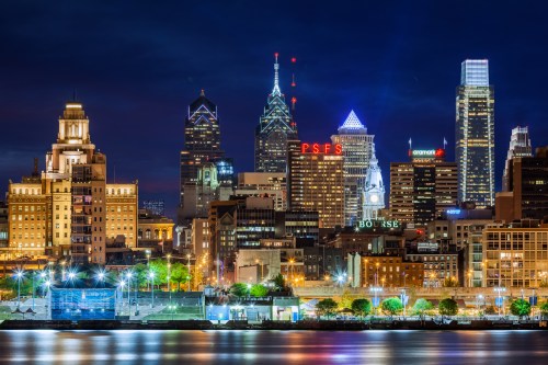 Philadelphia skyline at night, featuring illuminated skyscrapers and reflections on the water.