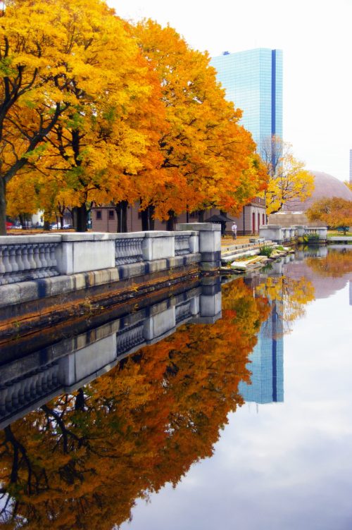 Autumn trees with vibrant orange leaves reflect in a calm river, alongside a stone wall and modern buildings.