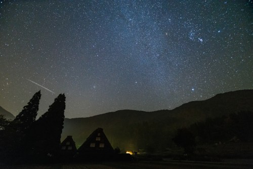 A starry night sky over mountains, with silhouettes of trees and a faint glow from a nearby building.