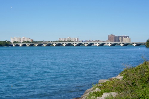 A wide bridge spans a calm blue river, with buildings lining the shore under a clear blue sky.
