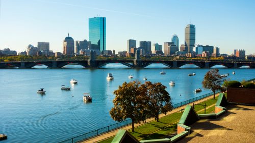 City skyline with tall buildings, a river, and boats, framed by trees and a bridge under a clear blue sky.