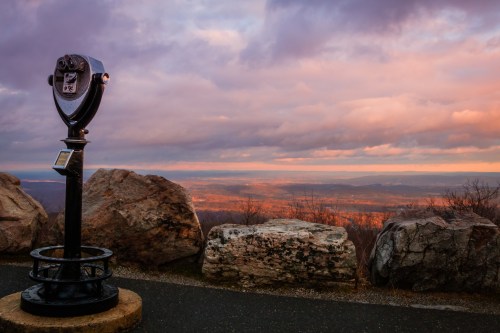 A scenic overlook with a viewing telescope, surrounded by rocks and a colorful sunset sky.