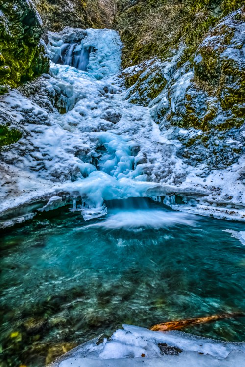A frozen waterfall cascades over rocky terrain into a turquoise pool, surrounded by icy formations and mossy rocks.