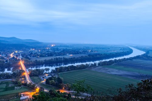 A scenic view of a river winding through fields and hills, illuminated by city lights at dusk.