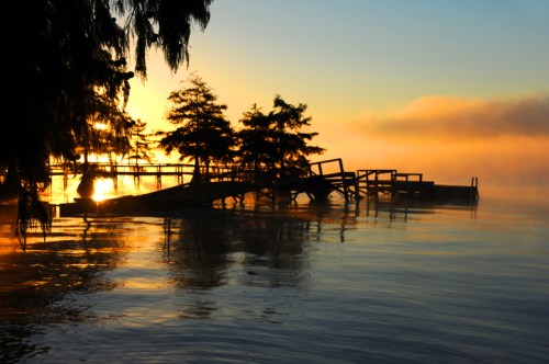 Sunrise over a misty lake, with silhouettes of trees and a wooden dock reflecting on the calm water.