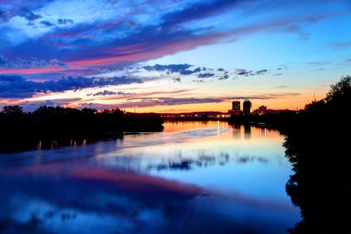 A serene river at sunset, reflecting vibrant colors in the sky and city skyline in the distance.