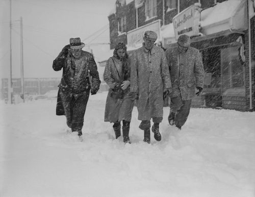 Four people walking through deep snow during a snowstorm, bundled in winter coats and hats, with shops in the background.