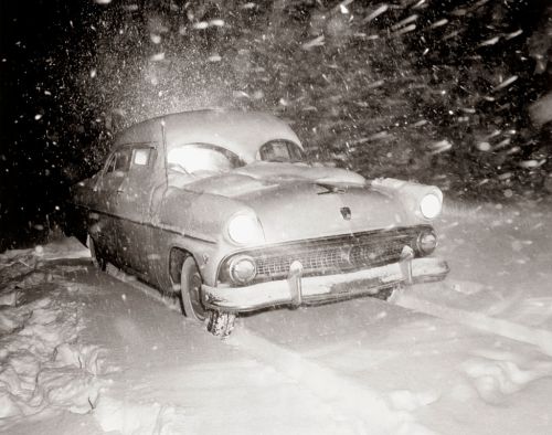 A vintage car partially buried in snow during a heavy snowstorm, with snowflakes swirling around it.