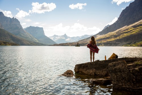 A person in a plaid shirt stands by a lake, surrounded by mountains and a clear sky, enjoying the serene landscape.