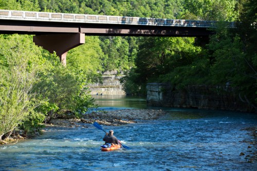 A person kayaking on a river beneath a bridge, surrounded by lush greenery and rocky banks.