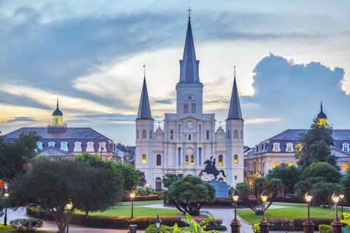 Historic church with spires at dusk, surrounded by trees and a statue, under a cloudy sky in a vibrant city.