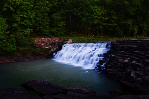 A serene waterfall cascades over rocky ledges into a calm, green pool, surrounded by lush trees.