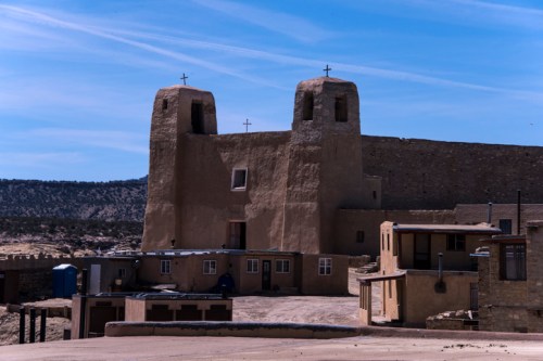 Historic adobe church with twin towers and crosses, surrounded by traditional buildings in a desert landscape.