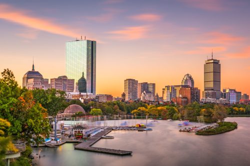 City skyline at sunset with water, boats, and trees in the foreground, showcasing modern buildings and a colorful sky.