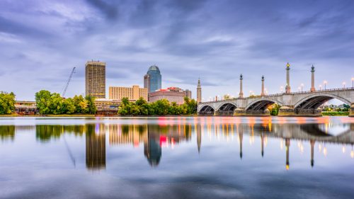 City skyline with modern buildings and a bridge reflected in a calm river under a cloudy sky at dusk.