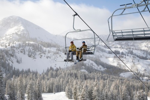 Two skiers on a chairlift, surrounded by snow-covered mountains and pine trees under a partly cloudy sky.
