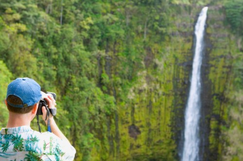 A person in a floral shirt takes a photo of a tall waterfall surrounded by lush greenery.