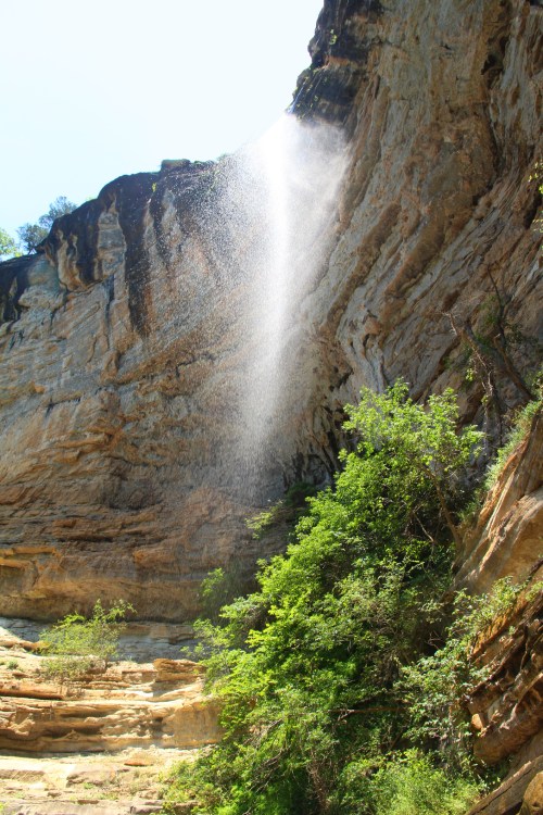 A waterfall cascades down a rocky cliff, surrounded by lush greenery and bright blue sky.