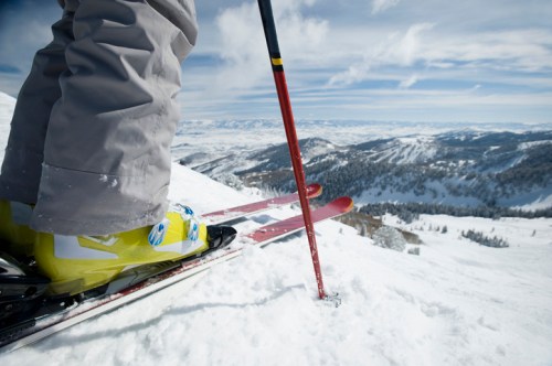 A skier's feet in bright yellow boots on skis, with a ski pole planted in the snow and a scenic mountain view in the background.
