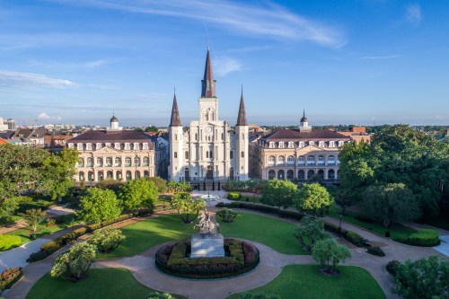 Aerial view of a historic building with spires, surrounded by lush gardens and a statue in a park. Clear blue sky above.