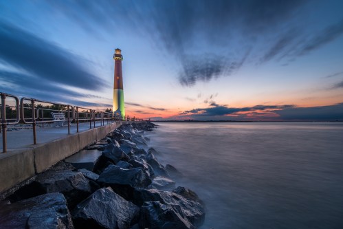 A lighthouse stands on a rocky pier at sunset, with colorful clouds reflecting in the calm water.