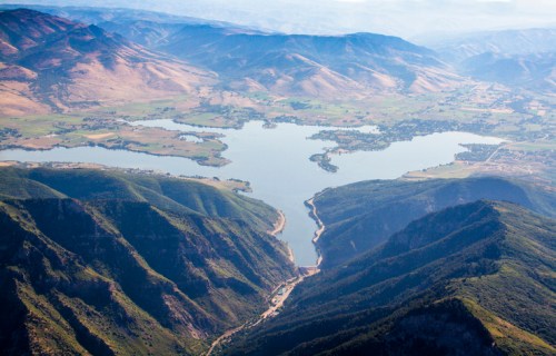 Aerial view of a serene lake surrounded by mountains and lush green valleys under a clear blue sky.