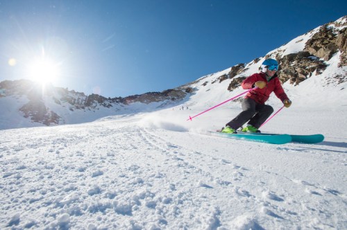 A skier in a red jacket glides down a snowy slope under a bright sun, with mountains in the background.