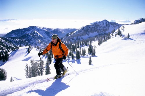 A skier in an orange jacket ascends a snowy mountain slope with trees and peaks in the background under a clear blue sky.