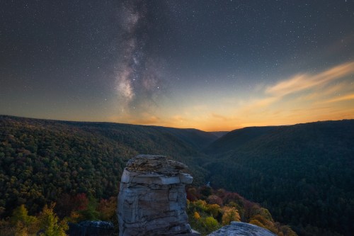 A stunning view of a valley at dusk, with a starry sky and the Milky Way above colorful autumn foliage.
