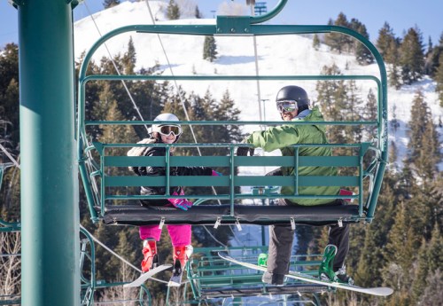 Two skiers on a chairlift, smiling and enjoying the snowy mountain scenery in the background.