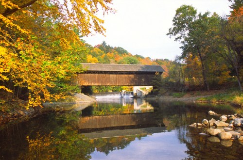 A covered bridge over a calm river, surrounded by vibrant autumn foliage and reflecting in the water.