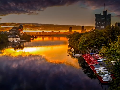 Sunset over a calm river, reflecting vibrant colors, with boats docked along the shore and city skyline in the background.